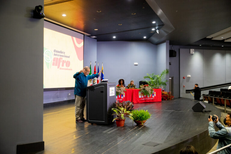 Persona frente a un podio declarando. Detraz hay una imagen proyectada que lee Cumbre Internacional Afro-desendencia