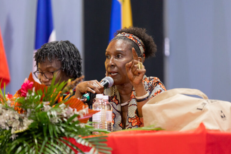 Dos mujeres afro-descendientes sentadas tras una mesa. La mujer de la derecha está hablando con micrófono en mano.