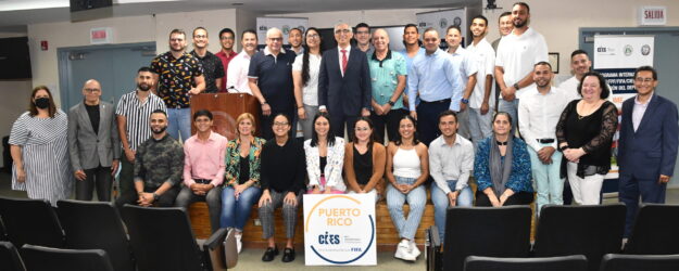 Segundo Cohorte Gestioìn del Deporte Dos filas de personas, las de atras estan trepadas en una tarima, las del frente sentadas en la tarima. De frente un afiche que lee Puerto Rico CIES, Red Universitaria Internacional. En colaboración con FIFA