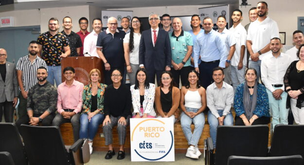 Dos filas de personas, las de atras estan trepadas en una tarima, las del frente sentadas en la tarima. De frente un afiche que lee Puerto Rico CIES, Red Universitaria Internacional. En colaboración con FIFA