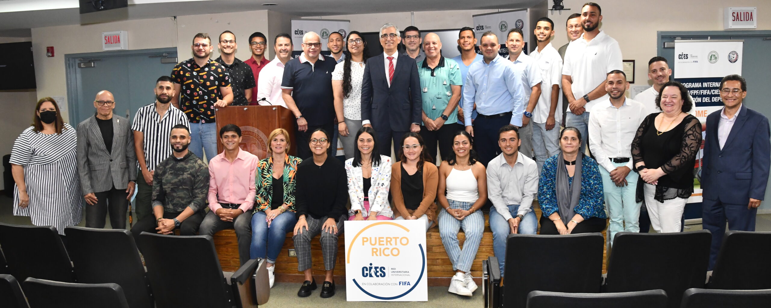 Segundo Cohorte Gestioìn del Deporte Dos filas de personas, las de atras estan trepadas en una tarima, las del frente sentadas en la tarima. De frente un afiche que lee Puerto Rico CIES, Red Universitaria Internacional. En colaboración con FIFA