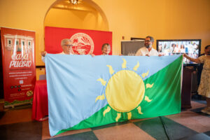 Alexis Massol, Angélica Varela y Arturo Massol soteniendo la bandera de Resiliencia energética. La bandera tiene en el centro un Sol color amarillo. Detraz del sol el fondo es dividido por la mitad desde la esquina inferior izquierda hasta la esquina superior derecha, formando dos triangulos. El triangulo de arriba es color azul claro, y el de abajo es color verde.