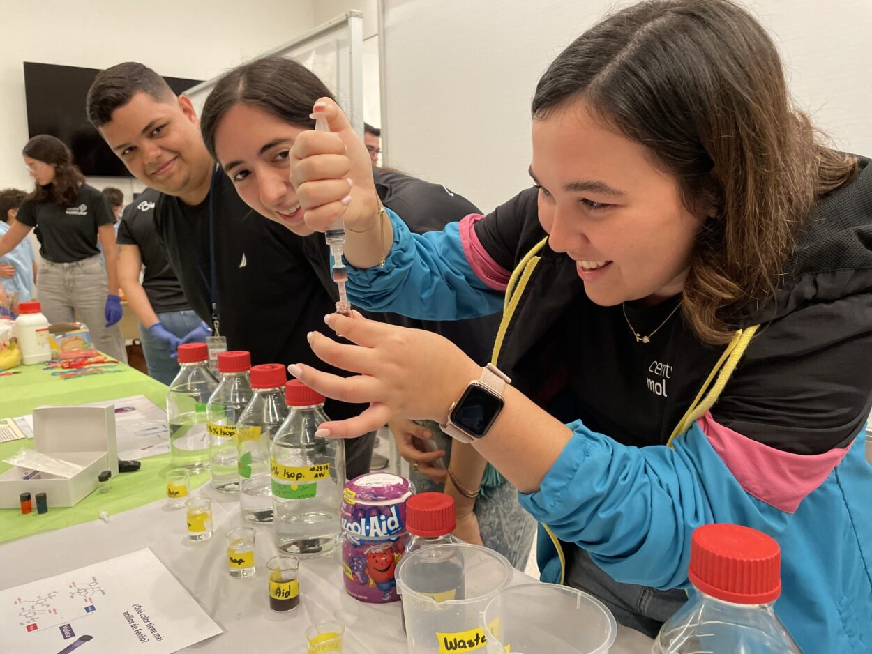 Estudiante virtiendo una sustancia en un envace que sostiene en la mano. Frente a la estudinte una mesa con envaces y botellas de plastico con diferentes sustancias.