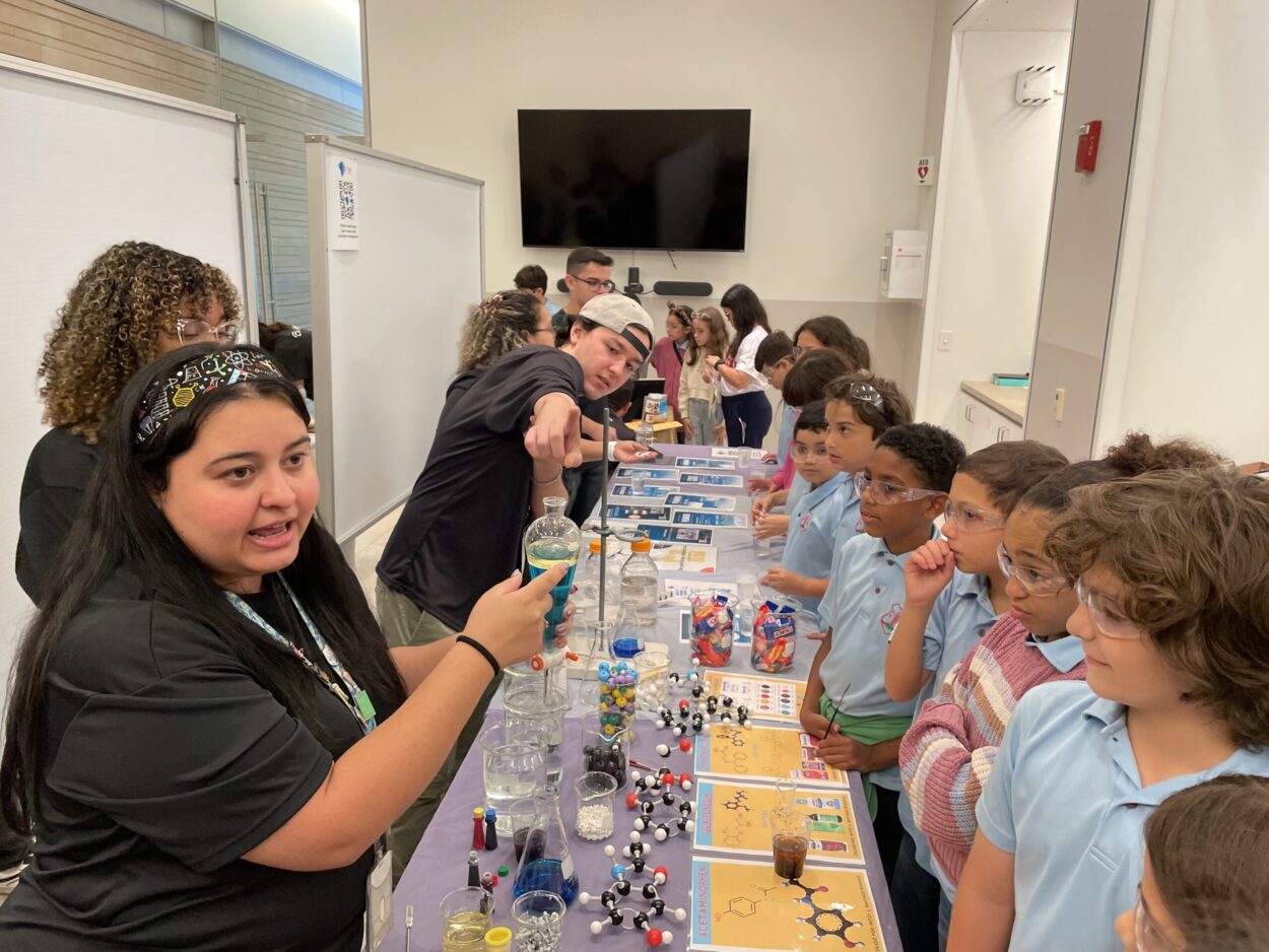 Una larga mesa angosta con materiales, sustancias, y hojas sueltas. A la izquierda de esta mesa están los estudiantes guías de Ciencias Naturales mostrando lo te está sobre la mesa. Y a la derecha de estan estudiantes de escuelas observando lo que muestran los guias.