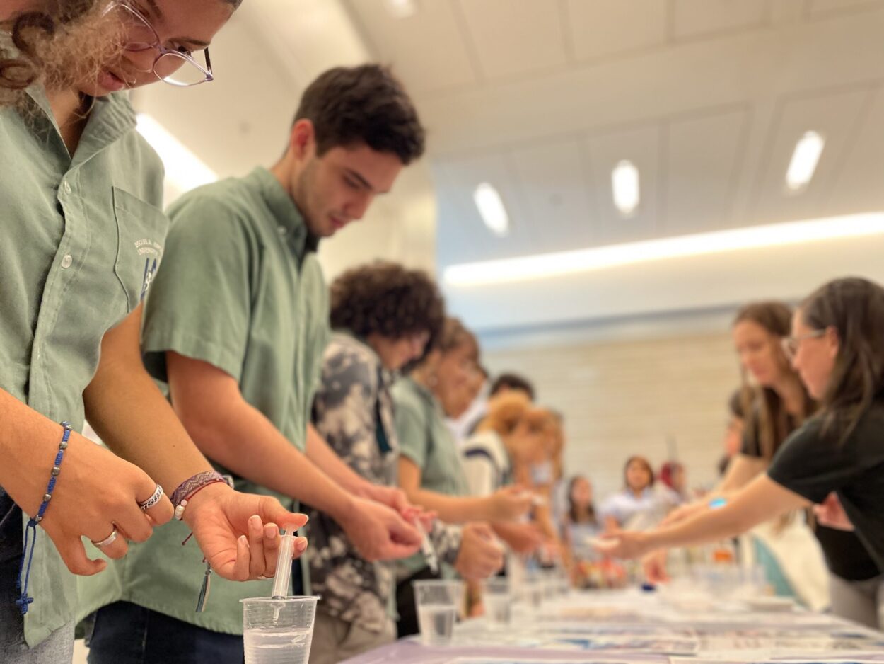 Acercamiento de estudiantes Guias de Ciencias Naturales trabajando con sustancias sobre una mesa. Frente a ellos unos estudiantes de escuela observando.