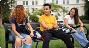 Tres estudiantes universitarios sentados en un banco al aire libre dentro del campus, conversando y compartiendo frente a un edificio universitario de UPR Cayey.