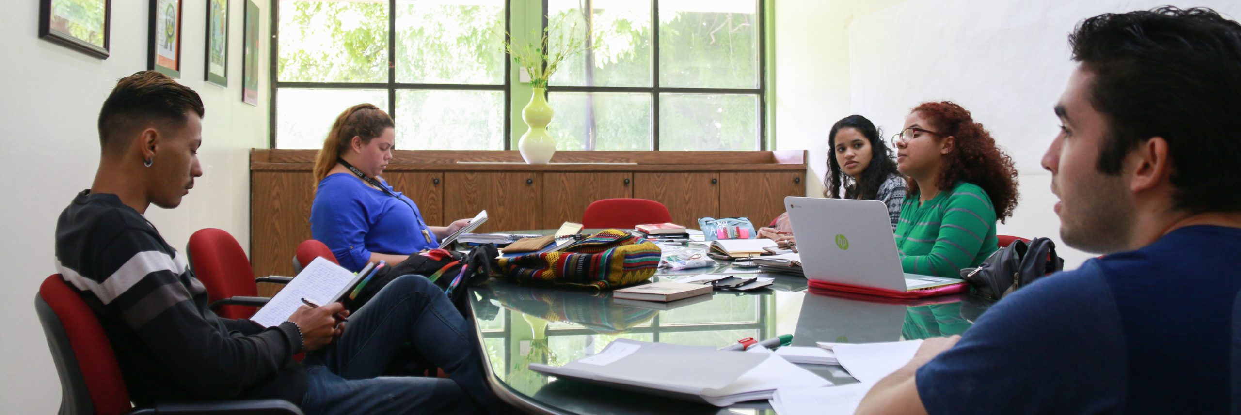 Cinco estudiantes en un salón sentados en una mesa grande. Algunos con libreta en mano escribiendo. Otro estudiante con una computadora portatil a su lado.