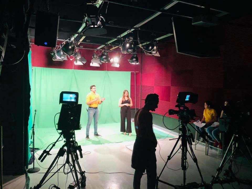 Un escenario con pantalla verde de fondo. Frente a la pantalla verde hay dos estudiantes que están siendo grabados por dos cámaras en primer plano junto con un camarografo.