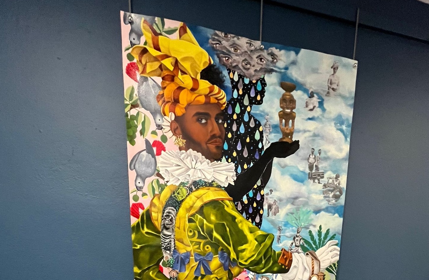 405429389_646094371069274_1820549664278499063_n Pintura abstracta. En primer plano un hombre de raza negra con turbante amarillo y lineas marron. Tiene un traje brillante color verde y amarillo, guante blanco en su mano derecha, y una cuellera frunsida. Su brazo izquierdo está totalmente negro, y sostiene una figura en la mano. El fondo del cuadro se divide en tres secciones verticales. La primera sección de la izquierda tiene unas cotorras grices posadas en una planta con frutos verdes y flores rojas. En el centro arriba una nube con muchos ojos, y debajo gotas de lluvias de diferentes colores en un fondo negro. A la derecha unas nubes con varias siluetas de personas de raza negra con vestido blanco que se mezcla con la nube.