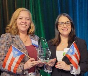 Dra. Moraima de Hoyos y la Lcda. Merién Méndez, sosteniendo la bandera de Puerto Rico y el premio a la excelencia en Innovación.