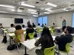 Salon con estudiantes sentados en pupitres.  De fondo se ve un instructor frente a una pizarra blanca.