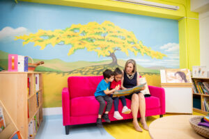 Niño y niña junto con una persona adulta sentados en un sofa rojo.  En el fondo un mural con un arbol.