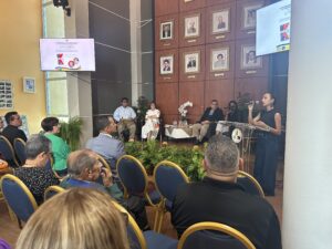 Vista de la ceremonia. Se ve el público sentado y al frente una persona hablando en un pódio. Bien al fondo se encuentra sentada la doctora Carmen Cazurro.