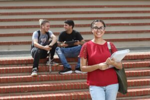 En el primer plano una estudiante frente a una escalera con su libreta en la mano. En el segundo plano dos estudiantes sentados en la escalera hablando entre sí.
