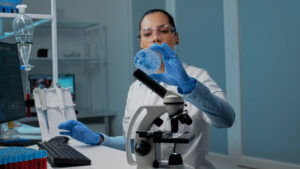 Chemistry scientist using research equipment in laboratory on desk with petri dish, micropipette, vacutainers and microscope. Scientific woman typing on computer for development