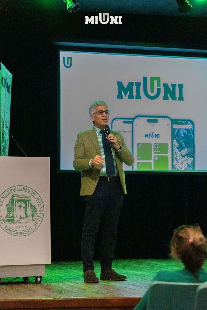 456084668_543479228240494_801685986044319780_n Moderador en podio frente a un afiche de la aplicación "Mi Uni"