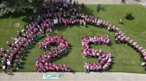 Foto aerea grupo de personas con camisas rosadas que están agrupadas de forma tal que están formando el número 25 dentro de un semicírculo.